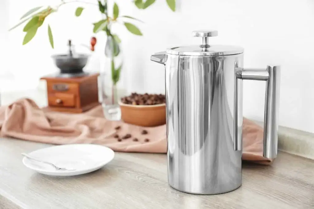 silver percolator on wooden counter next to white plate and coffee beans