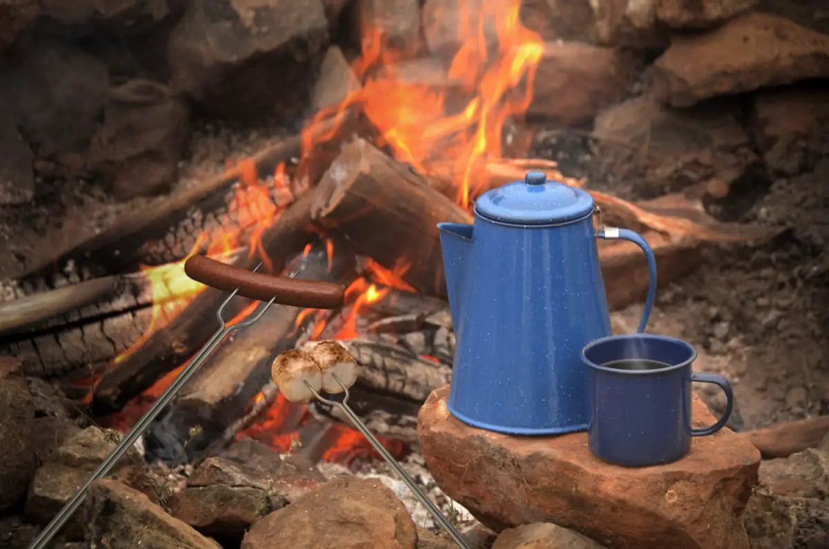 blue percolator by campfire beside cup of coffee, roasted marshmallows, and hotdogs