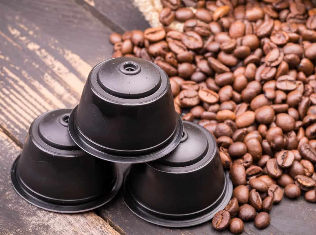 Three Nespresso capsules on a wooden table with a spilled bag of whole coffee beans beside them.