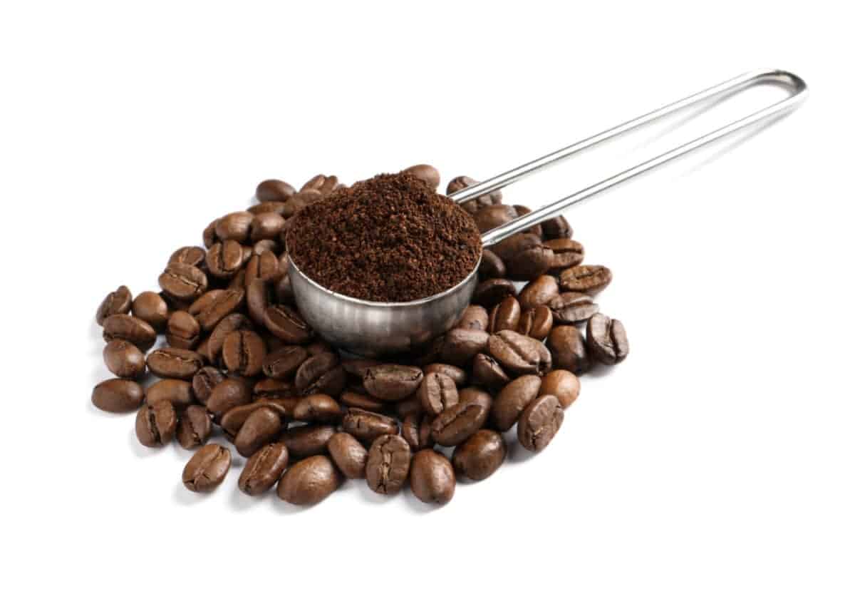 Scoop of ground coffee sitting on pile of coffee beans on white background.