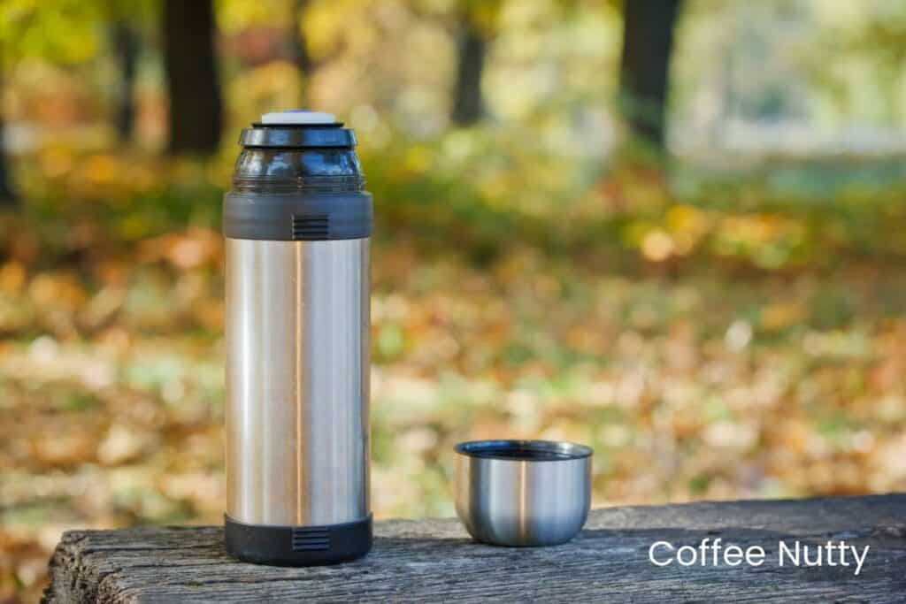 Coffee thermos with lid off, sitting on wooden log outdoors.