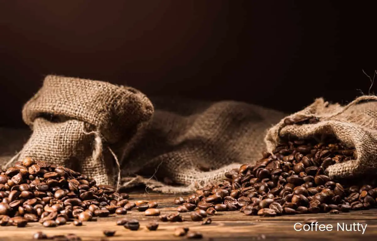Coffee beans spilling out of several burlap bags on wood table with dark backdrop.