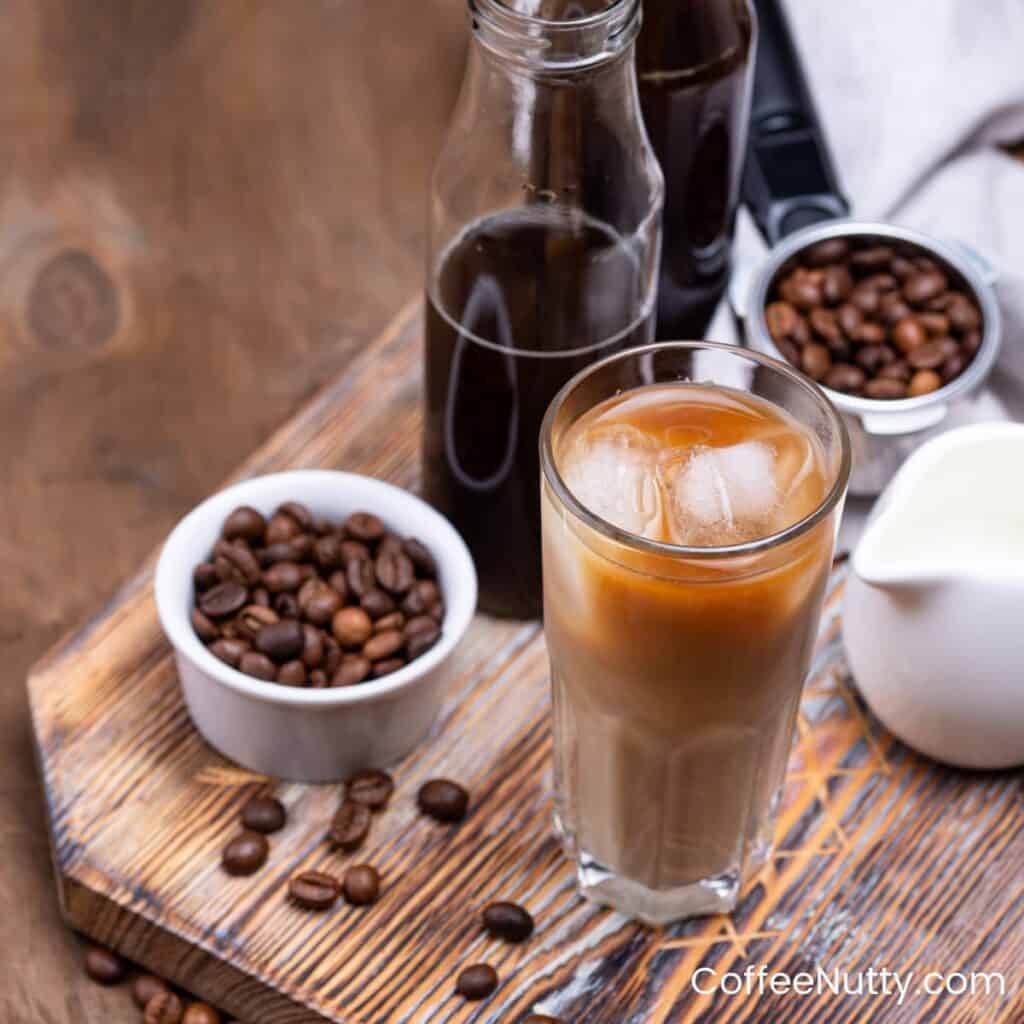 Cold brew with ice and milk in clear glass on wooden table next to coffee beans.