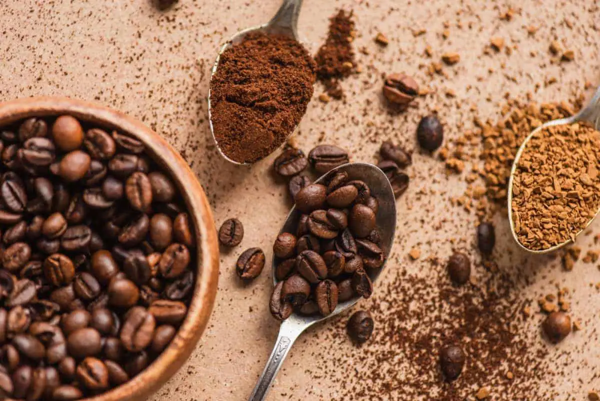 Whole beans in a bowl and spoon beside instant coffee and ground coffee in spoons.