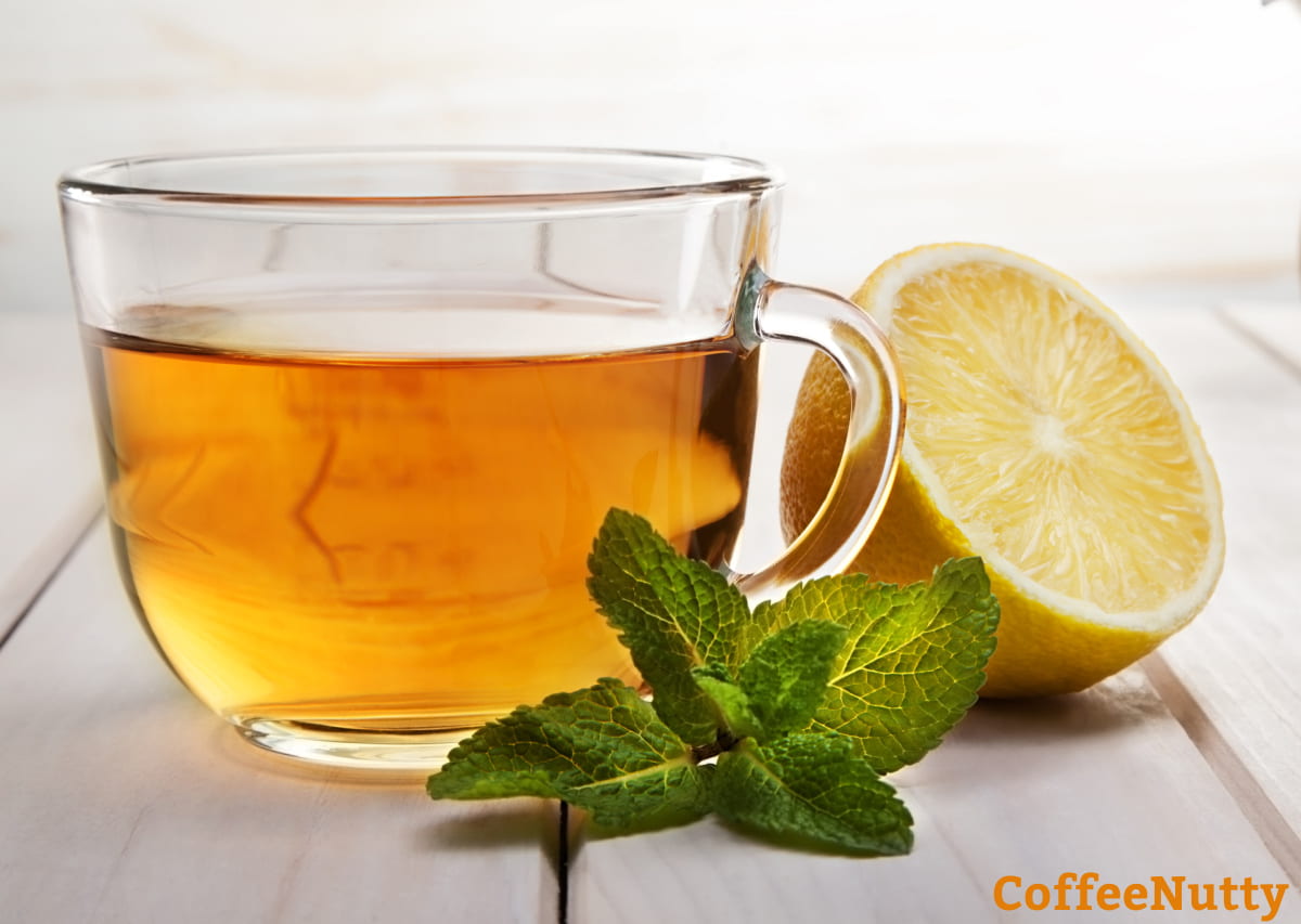 Cup of brewed tea in clear glass on light wood table by lemon and mint leaves.