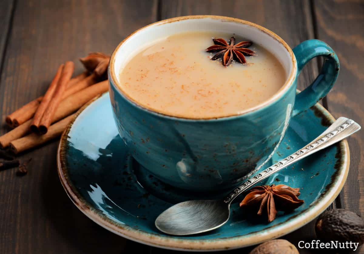 Chai tea with milk in cup with spoon on saucer to stir surrounded my spices including cinnamon sticks.